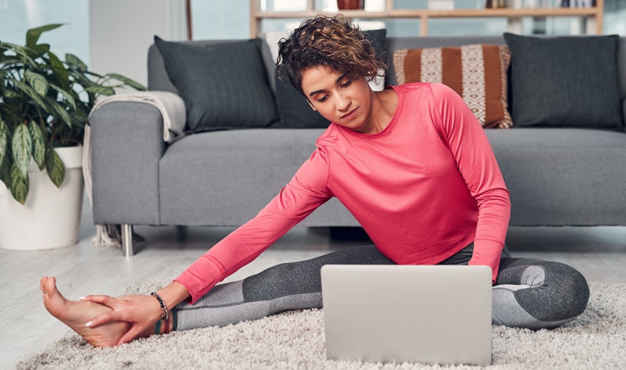 A woman does yoga in her living room while watching a laptop, placed on the floor.